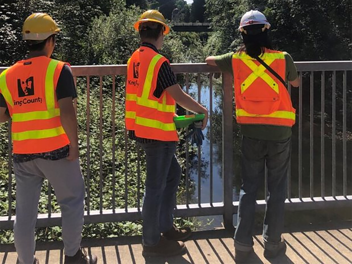 Interns and a bridge engineer looking over a bridge railing at the water below.