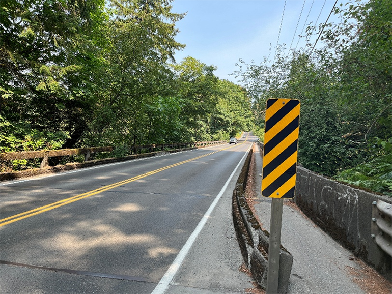 Looking down Judd Creek Bridge from the sidewalk on a sunny day with Blue skies, a striped hazard sign on the right.