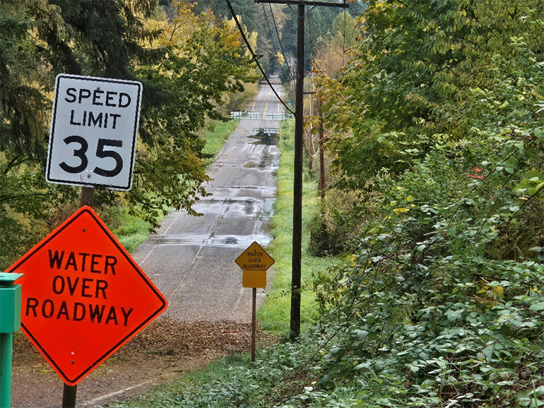 Orange water over roadway sign in front of NE 179th Place, water can be seen over the road. 