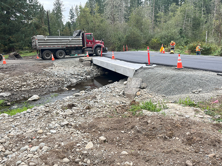 A large truck drives over a road with fresh smooth asphalt above the new concrete box culvert. 