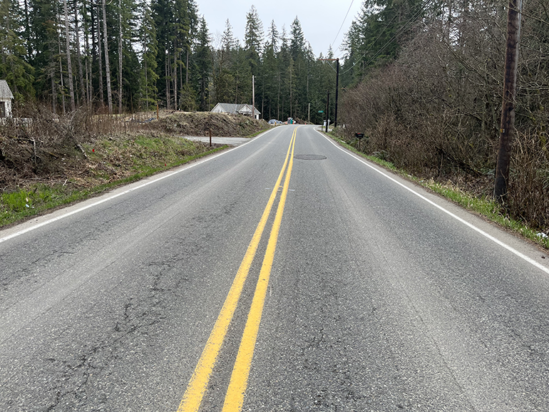 Straight road with yellow stripes down the middle showing cracks in the pavement and patching over potholes.