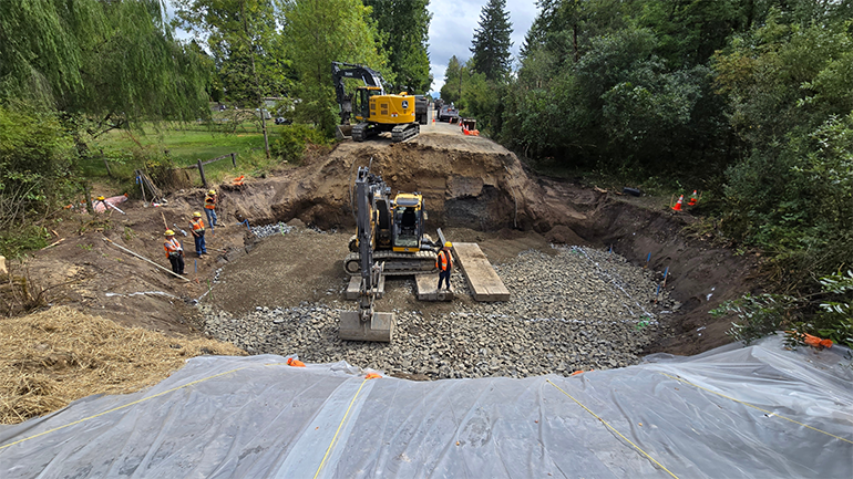 Crew members and escavators in a deep pit in the road as they prepare for the upcoming placement of the large concrete box culvert by crane.