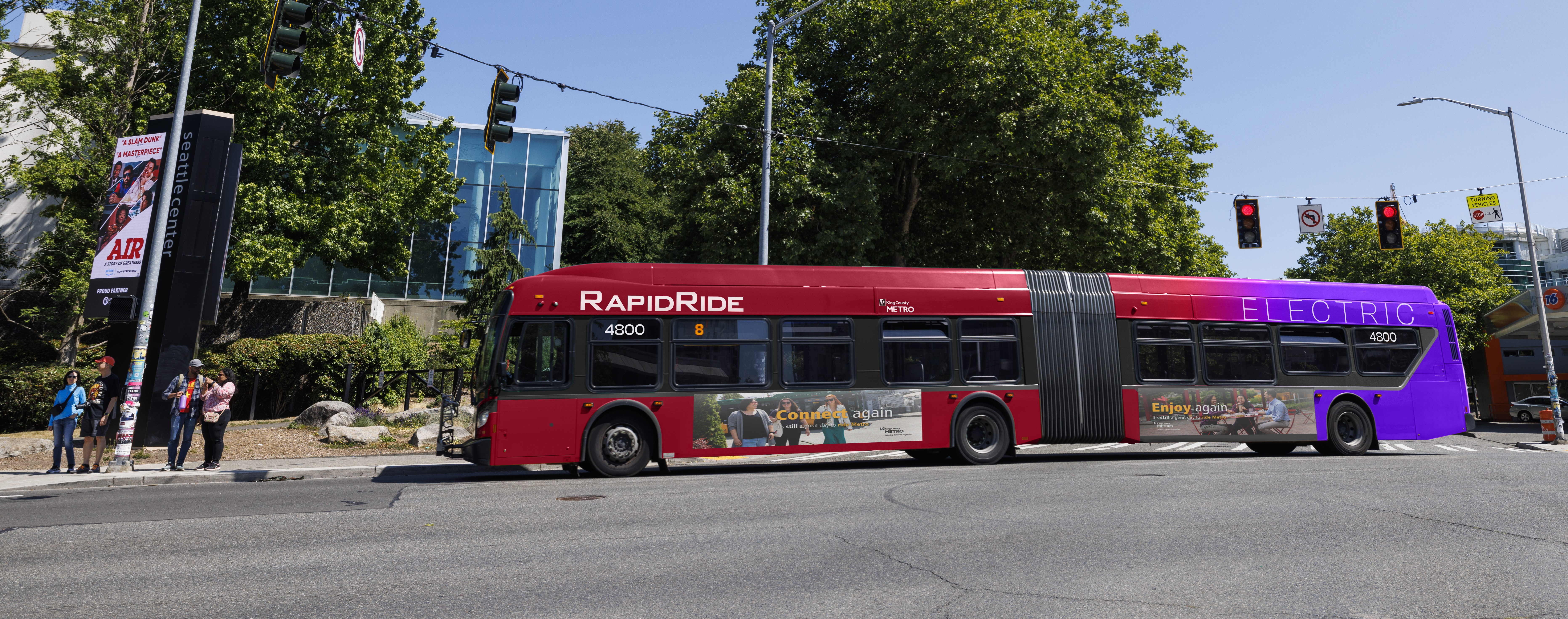 Rendering of a Metro RapidRide bus wrapped in the new red and purple design on tree-lines street against a blue sky
