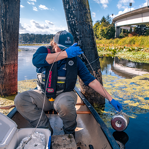 A person wearing safety gear sits in a canoe and lowers a water quality testing device into the water.