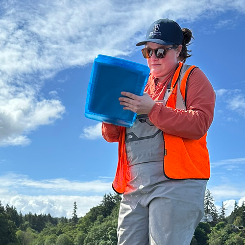 A person wearing waders, an orange safety vest, and a King County hat makes notes on a blue clipboard.