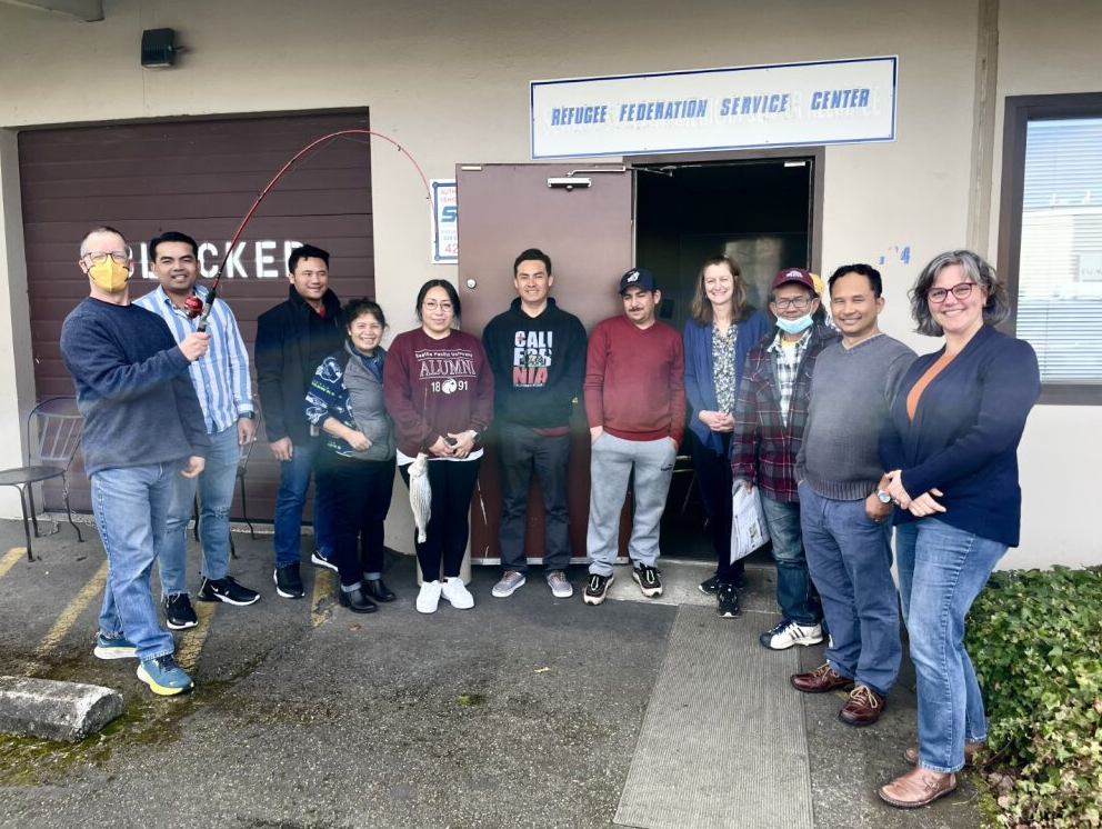 11 people stand in a line smiling. One of them holds a fish on a fishing pole. Above them is a sign that reads "Refugee Federation Service Center."