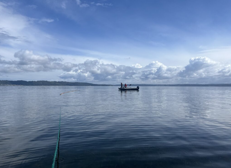 A wide shot of water and sky in Puget Sound. A green rope trails into the distance toward a small boat with two people visible aboard.